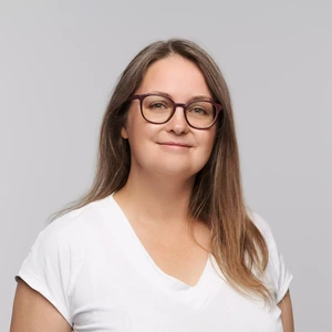 Portretinė nuotrauka – jauna moteris ilgais, tiesiais šviesiai rudais plaukais, dėvinti baltus marškinėlius ir akinius. Ji žiūri tiesiai į kamerą neutraliu žvilgsniu. Fonas neutralus, šviesiai pilkas.
