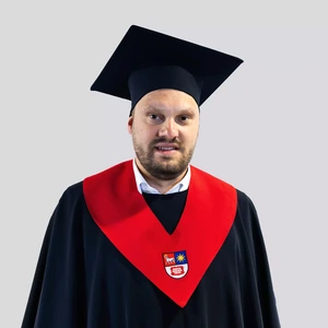 Portretinė nuotrauka – vyras apsirengęs akademiniais drabužiais: tamsia mantija su ryškiai raudonu apykaklės akcentu ir herbu, bei tamsia akademine kepure. Jis žvelgia į kamerą rimta išraiška. Fonas neutralus, šviesiai pilkas.