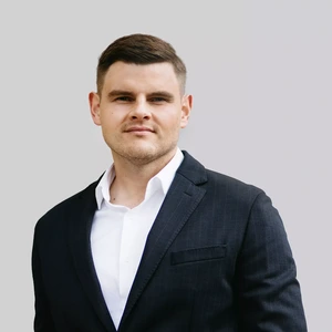 Portretinė nuotrauka – vyras trumpais šviesiais plaukais, vilkintis tamsų švarką ir baltus marškinius. Jis žiūri į kamerą rimtu žvilgsniu, fonas neutralus, pilkšvas.