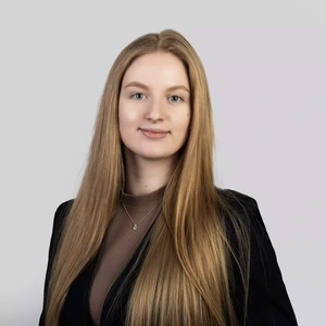 Portretinė nuotrauka – moteris su ilgais šviesiais plaukais, vilkinti tamsiai mėlyną švarką ir smėlio spalvos palaidinę. Ji šypsosi ir žiūri į kamerą, fone pilka spalva.