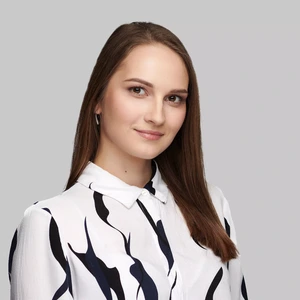 Jauna moteris tiesiais rudais plaukais dėvi baltus marškinius su juodais ornamentiniais raštais, žiūri į kamerą švelniai šypsodamasi šviesiame fone.