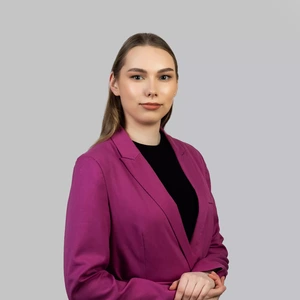 Portretinė nuotrauka – jauna moteris tiesiais šviesiais plaukais, dėvinti ryškiai fuksijos spalvos švarką ir juodą palaidinę. Ji žiūri tiesiai į kamerą rimtu, pasitikinčiu žvilgsniu, rankos laikomos priešais. Fonas neutralus, šviesiai pilkas.