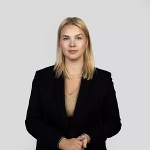 Portretinė nuotrauka – jauna moteris šviesiais tiesiais plaukais, dėvinti juodą švarką ir smėlio spalvos palaidinę. Ji stovi tiesiai, žiūri į kamerą rimtu, bet ramiu žvilgsniu. Fonas šviesiai pilkas, neutralus.