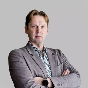 Vyresnio amžiaus vyras su šviesiais plaukais, vilkintis rusvą švarką ir raštuotus marškinius, stovi sukryžiavęs rankas ir rimtu veidu žiūri į kamerą, fonas šviesiai pilkas.