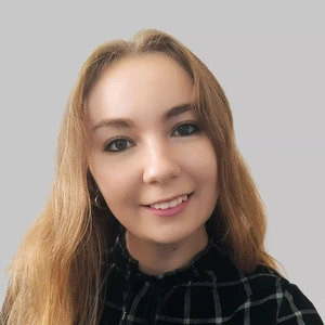 Jauna moteris su šviesiais, pusilgiais plaukais, vilkinti juodus languotus marškinius, šypsosi ir žiūri į kamerą, fonas pilkas.
