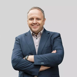 Vidutinio amžiaus vyras trumpais šviesiais plaukais, vilkintis tamsiai mėlynu kostiumu ir šviesiais marškiniais, susidėjęs rankas ir besišypsantis, fonas pilkas.