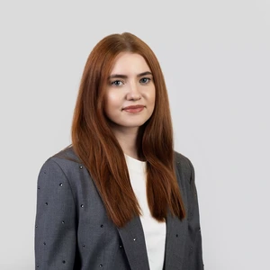 Portretinė nuotrauka – moteris ilgais tiesiais vario raudonumo plaukais, vilkinti pilku švarku su dekoratyvinėmis detalėmis ir baltais marškinėliais. Ji žiūri į kamerą su lengva šypsena. Fonas – šviesiai pilkas.