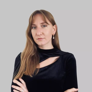 Rimta moteris ilgais šviesiai rudais plaukais, vilkinti juodu aksominiu viršumi su iškirptu akcentu, dėvinti perlo tipo auskarus, pilkas fonas.