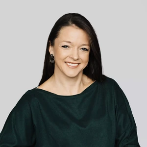Portretinė nuotrauka – moteris ilgais tamsiais plaukais, vilkinti tamsiai žalią palaidinę. Ji šypsosi ir žiūri į kamerą neutraliame pilkame fone. Ausyse – elegantiški juodi auskarai su pakabukais.