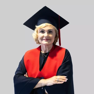 Portretinė nuotrauka – moteris šviesiais garbanotais plaukais, vilkinti akademinę mantiją su ryškiai raudonu apykaklės akcentu ir tamsia akademine kepure su raudonu kutu. Ji stovi sukryžiavusi rankas ir šypsosi žiūrėdama į kamerą. Fonas neutralus, šviesiai pilkas.