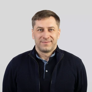 Portretinė nuotrauka – vyras trumpais šviesiais plaukais, vilkintis tamsiai mėlyną megztinį su užtrauktuku ir šviesius marškinius. Jis žiūri į kamerą su švelnia šypsena, fonas neutralus, pilkšvas.