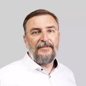 Portretinė nuotrauka – vyresnis vyras su pilna barzda, vilkintis baltus marškinius. Jis žiūri į kamerą rimta veido išraiška, fonas neutralus, pilkšvas.