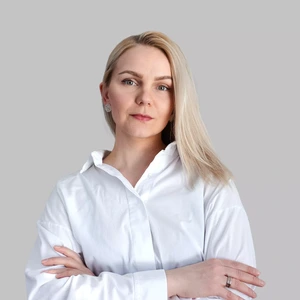 Moteris šviesiais plaukais dėvi baltus marškinius, stovi sukryžiavusi rankas ir žvelgia į kamerą rimtu, pasitikinčiu žvilgsniu šviesiame fone.