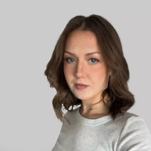 Portretinė nuotrauka – jaunas vyras šviesiais plaukais, sušukuotais atgal, vilkintis šviesiai mėlynus marškinius ir tamsų megztinį. Jis šypsosi ir žvelgia tiesiai į kamerą. Fonas neutralus, pilkšvas.