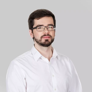 Rimtai atrodantis vyras trumpais tamsiais plaukais ir barzda, vilkintis baltus marškinius, dėvintis akinius, pilkas fonas.