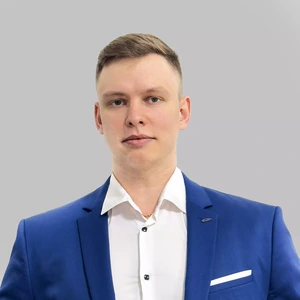 Jaunas vyras trumpais šviesiais plaukais, vilkintis ryškiai mėlynu kostiumu ir baltais marškiniais, žiūri tiesiai į kamerą, fonas pilkas.