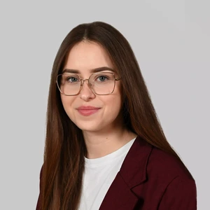 Portretinė nuotrauka – jauna moteris ilgais, tiesiais tamsiai rudais plaukais. Ji dėvi akinius, baltus marškinėlius ir tamsiai raudoną švarką. Fonas – šviesiai pilkas.