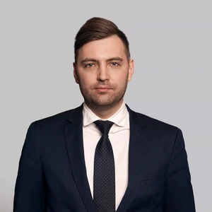 Portretinė nuotrauka – vyras trumpais plaukais, vilkintis tamsų kostiumą su baltais marškiniais ir tamsiu taškuotu kaklaraiščiu. Jis žiūri į kamerą rimta veido išraiška, fonas neutralus, pilkšvas.
