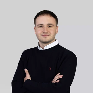 Portretinė nuotrauka – jaunas vyras trumpais tamsiais plaukais, dėvintis baltus marškinius ir tamsiai mėlyną megztinį su nedideliu raudonu simboliu priekyje. Jis stovi sukryžiavęs rankas ir žiūri į kamerą su lengva šypsena. Fonas neutralus, šviesiai pilkas.