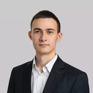 Portretinė nuotrauka – jaunas vyras trumpais tamsiais plaukais, dėvintis tamsų švarką ir baltus marškinius. Jis žiūri į kamerą rimtu, neutraliu žvilgsniu. Fonas neutralus, šviesiai pilkas.