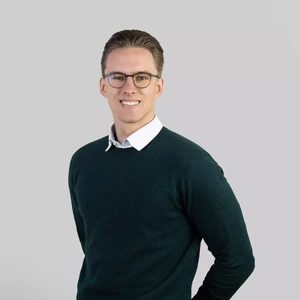 Portretinė nuotrauka – jaunas vyras trumpais šviesiais plaukais, vilkintis tamsiai žalią megztinį su baltais marškiniais, nešiojantis akinius. Jis žiūri į kamerą su plačia šypsena, fonas neutralus, pilkšvas.