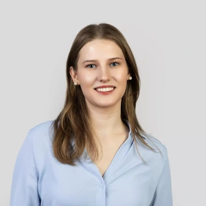 Portretinė nuotrauka – jauna moteris tiesiais šviesiai rudos spalvos plaukais, dėvinti šviesiai mėlynus marškinius ir perlinius auskarus. Ji šypsosi žiūrėdama į kamerą, fonas šviesus ir neutralus.