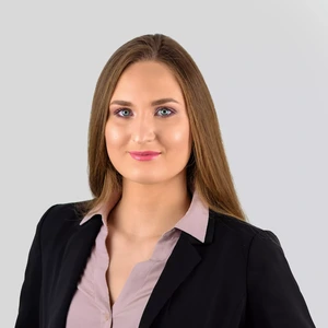 Portretinė nuotrauka – jauna moteris ilgais, šviesiai rudais plaukais. Ji dėvi šviesiai rausvus marškinius ir juodą švarką. Fonas – šviesiai pilkas.