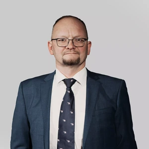 Vidutinio amžiaus vyras su akiniais ir barzdele dėvi tamsiai mėlyną kostiumą, baltus marškinius ir tamsų kaklaraištį su mažais gyvūnų motyvais. Jis žiūri tiesiai į kamerą rimtu veidu šviesiame fone.