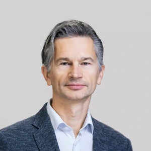 Portretinė nuotrauka – vidutinio amžiaus vyras su žilstelėjusiais plaukais, vilkintis pilkšvu švarku ir šviesiais marškiniais, žiūri tiesiai į kamerą neutraliame pilkame fone.