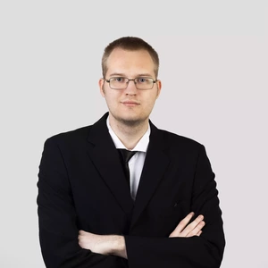 Vyras trumpais plaukais ir akiniais, vilkintis juodą kostiumą, stovi sukryžiavęs rankas prieš šviesų foną