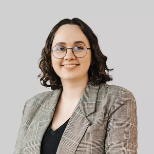 Portretinė nuotrauka – jauna moteris trumpais garbanotais plaukais, dėvinti languotą švarką ir juodą palaidinę. Ji nešioja apvalius akinius su plonais rėmeliais ir šypsosi žiūrėdama į kamerą. Fonas neutralus, šviesiai pilkas.