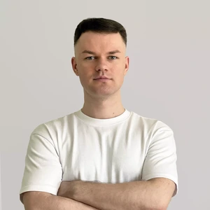 Portretinė nuotrauka – jaunas vyras trumpais tamsiais plaukais, dėvintis paprastus baltus marškinėlius. Jis stovi sukryžiavęs rankas ir žiūri į kamerą rimtu žvilgsniu. Fonas neutralus, šviesiai pilkas.
