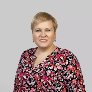Portretinė nuotrauka – moteris trumpais šviesiais plaukais, dėvinti spalvingą raštuotą palaidinę su rožinių ir raudonų gėlių motyvais. Ji žvelgia į kamerą su malonia šypsena. Fonas neutralus, šviesiai pilkas.