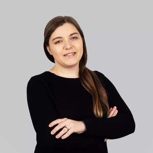 Portretinė nuotrauka – moteris ilgais šviesiai rudais plaukais, vilkinti juodus drabužius, šypsosi žvelgdama į kamerą šviesiame fone. Rankos sukryžiuotos, matyti vestuvinis žiedas.