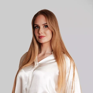Jauna moteris ilgais šviesiais plaukais dėvi baltą šilkinę palaidinę, žvelgia tiesiai į kamerą šviesiame fone su ramiu, pasitikinčiu žvilgsniu.