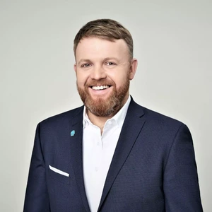 Portretinė nuotrauka – vyras su trumpais šviesiais plaukais ir pilnu barzda, vilkintis tamsiai mėlyną kostiumą su baltais marškiniais. Jis šypsosi ir žiūri į kamerą, fone šviesi spalva.