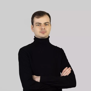 Portretinė nuotrauka – jaunas vyras trumpais plaukais, vilkintis juodą golfą. Jis stovi sukryžiavęs rankas ir žiūri tiesiai į kamerą neutraliu veido išraiška. Fonas šviesiai pilkas, neutralus.