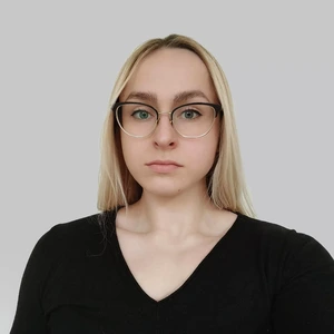 Portretinė nuotrauka – jauna moteris šviesiais plaukais, su akiniais ir juodais marškiniais, žvelgia tiesiai į kamerą šviesiame fone.