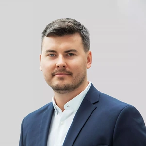 Portretinė nuotrauka – vyras trumpais plaukais ir šviesia barzda, vilkintis baltus marškinius bei tamsiai mėlyną švarką. Jis žvelgia tiesiai į kamerą su lengva šypsena. Fonas – šviesiai pilkas.