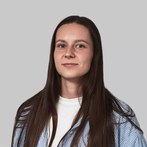 Portretinė nuotrauka – jauna moteris ilgais tamsiais plaukais, dėvinti baltus marškinėlius ir dryžuotus šviesiai mėlynai baltus marškinius. Ji žiūri į kamerą ramia išraiška. Fonas neutralus, šviesiai pilkas.