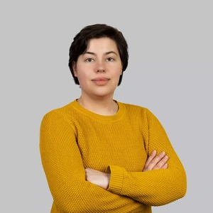 Portretinė nuotrauka – moteris trumpais tamsiais plaukais, dėvinti garstyčių geltonumo megztinį. Ji stovi sukryžiavusi rankas ir žiūri į kamerą rimtu, ramiu žvilgsniu. Fonas neutralus, šviesiai pilkas.