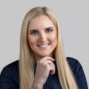 Moteris ilgais šviesiais plaukais dėvi tamsius marškinius, šypsosi ir švelniai remiasi smakru į ranką, žvelgdama į kamerą šviesiame fone.