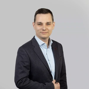 Portretinė nuotrauka – jaunas vyras trumpais tamsiais plaukais, dėvintis šviesiai mėlynus marškinius ir tamsų švarką su segtuku atlapuose. Jis žvelgia į kamerą su lengva šypsena, fonas šviesus, neutralus.