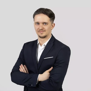 Portretinė nuotrauka – vyras trumpais plaukais, vilkintis tamsų kostiumą ir baltus marškinius. Jis stovi sukryžiavęs rankas ir žiūri į kamerą, fonas neutralus, pilkšvas.