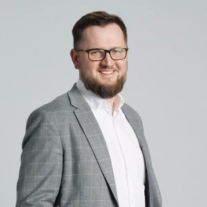 Portretinė nuotrauka – vyras su trumpais plaukais ir pilna barzda, vilkintis šviesiai pilką kostiumą su baltais marškiniais. Jis šypsosi ir žiūri į kamerą, fone šviesi spalva.