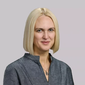 Šviesiaplaukė moteris tiesiais plaukais, vilkinti pilką palaidinę su stovu, žiūri į kamerą ir šypsosi.