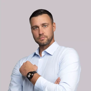Rimtos išraiškos vyras trumpais tamsiais plaukais, dėvintis šviesiai mėlynus marškinius ir juodą laikrodį, pozuoja šviesiame fone sukryžiuotomis rankomis.