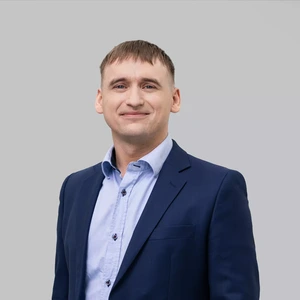Portretinė nuotrauka – jaunas vyras su trumpai kirptais plaukais, vilkintis tamsiai mėlynu kostiumu ir šviesiai mėlynais marškiniais, šypsosi žvelgdamas į kamerą šviesiame fone.