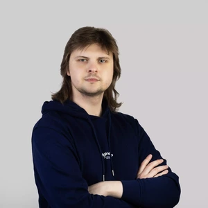 Portretinė nuotrauka – jaunas vyras pusilgiais plaukais, vilkintis tamsiai mėlyną džemperį su gobtuvu. Jis stovi sukryžiavęs rankas, žvelgia į kamerą rimtu žvilgsniu. Fonas neutralus, šviesiai pilkas.