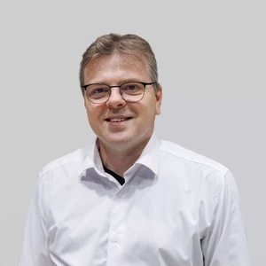 Vyras su akiniais ir trumpais plaukais, vilkintis baltus marškinius, žiūri į kamerą ir šypsosi.